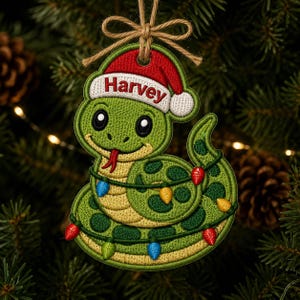 May include: An embroidered ornament shaped like a green snake with dark green spots, wearing a red and white Santa hat with the name Harvey embroidered on the brim. The snake is wrapped in a string of colorful Christmas lights and hangs by twine.