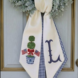 May include: Cream-colored door scarf with a blue and white gingham trim. One side features an embroidered topiary with American flags and a red bow. The other side has a navy blue embroidered letter "L".