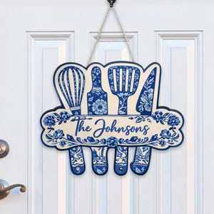 Puede incluir: Un letrero de cocina de madera con un diseño floral azul y blanco. El letrero presenta un batidor, una espátula, un cuchillo y otros utensilios de cocina. El texto "The Johnsons" se muestra en una fuente decorativa.
