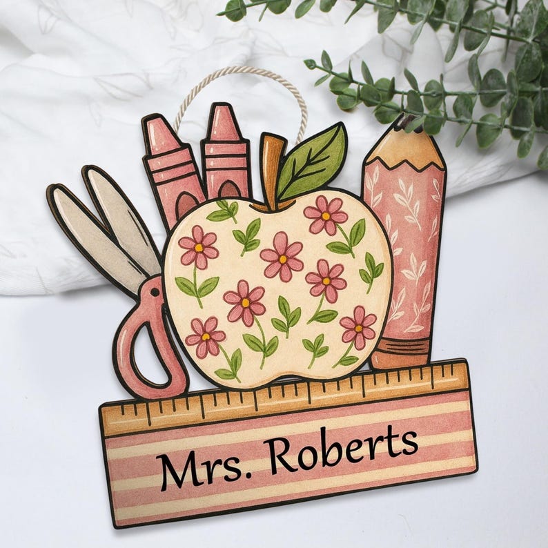 May include: Wooden teacher-themed ornament featuring an apple with pink flowers, a pencil, crayons, and scissors. A ruler at the bottom has the name "Mrs. Roberts" printed on it. The ornament is designed with a neutral color palette.