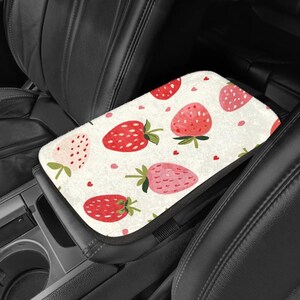 May include: A car armrest cover featuring a repeating pattern of red and pink strawberries with green leaves and small pink hearts on a cream background. The cover is rectangular and fits over the center console armrest.