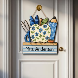 May include: A wooden teacher's door sign featuring an apple with blue flower details, scissors, crayons, and a pencil. The sign includes a ruler with the name "Mrs. Anderson" and is hanging on a white door.