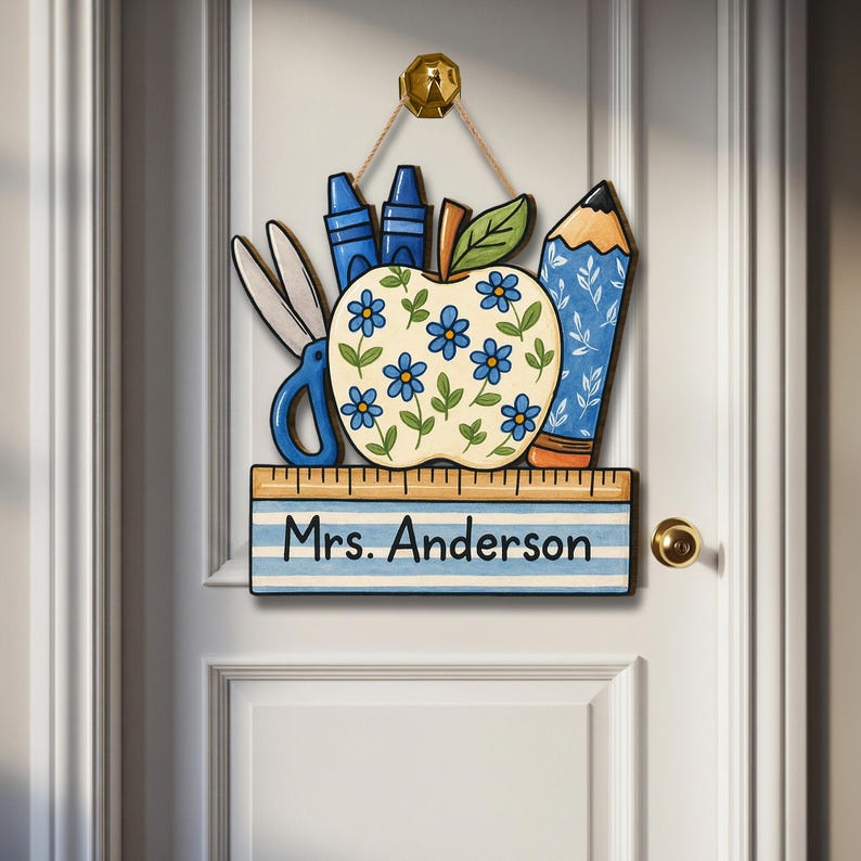 May include: A wooden teacher's door sign featuring an apple with blue flower details, scissors, crayons, and a pencil. The sign includes a ruler with the name "Mrs. Anderson" and is hanging on a white door.