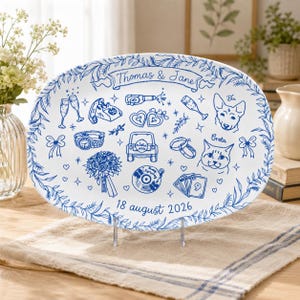 Könnte beinhalten: Eine ovale weiße Keramikplatte mit blauen Strichzeichnungen von Hochzeitssymbolen wie Champagnergläsern, einem Blumenstrauß, Ringen und Haustieren. Der Text lautet "Thomas & Jane" sowie "18 august 2026" mit einem Blatt-Rand.