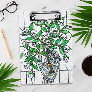 May include: A decorative clipboard with a stained-glass design. It features a tree of skulls and green leaves, held by a metallic robotic hand. The clipboard has a silver metal clip at the top.