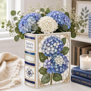 Könnte beinhalten: Eine rechteckige Keramikvase in Buchform mit dem Text Taylor's Library auf dem Buchrücken. Die Vase hat einen weißen Hintergrund mit blauen und weißen Hortensien-Illustrationen und Goldrand, gefüllt mit blauen und weißen Hortensien.