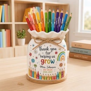 May include: A white ceramic pencil holder with a scalloped rim and a jute twine bow. The front features colorful illustrations of flowers, a rainbow, and pencils, with the text Thank you for helping us grow, Mrs. Johnson, 3rd Grade, 2026-2027.