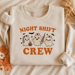 May include: Cream-colored sweatshirt with the words "NIGHT SHIFT CREW" in orange block letters. The design features three cartoon ghost characters wearing nurse hats, with a pumpkin and syringe. Black bats and stars complete the design.