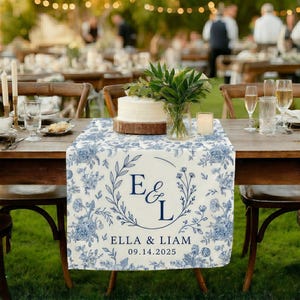 Puede incluir: Una mesa de boda con un camino de mesa con estampado floral azul y blanco. El camino de mesa presenta las iniciales "E & L" dentro de una corona, junto con los nombres "ELLA & LIAM" y la fecha "09.14.2025". Una tarta blanca se encuentra sobre una base de madera.