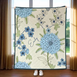 May include: A cream-colored quilt featuring a floral design with large blue flowers and smaller blue blossoms. The quilt has a dark green border and the name "Emily" is written in cursive. The quilt is displayed in front of a window.