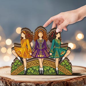 May include: Wooden Irish dancers figurine. Three figures in colorful dresses of yellow, purple, and green, with Celtic knot designs. The base features shamrocks and more Celtic patterns. The figurine is on a wooden surface.