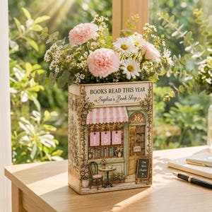 Puede incluir: Un florero rectangular con forma de libro que presenta una ilustración impresa de un escaparate con la etiqueta 'BOOKS READ THIS YEAR' y 'Sophia's Book Shop' con el número 158, lleno de claveles rosas, margaritas blancas y eucalipto verde.