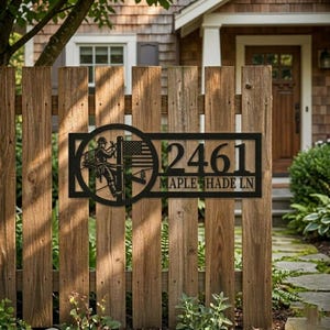 May include: Black metal address sign with the number 2461 and "MAPLE SHADE LN" below it. The sign features a circular design with a lineman and an American flag. The sign is mounted on a weathered wooden fence.