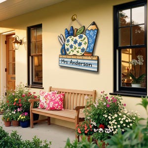 May include: A wooden teacher's sign with an apple, pencil, scissors, and crayons design. The sign is painted with blue and white stripes and features the name "Mrs. Anderson". The sign is hanging on a beige wall near a wooden bench and flower pots.