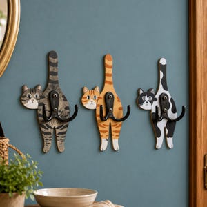 May include: Three wall-mounted hooks shaped like cat bodies with tails pointing upward. Each hook features a different cat pattern: gray tabby, orange tabby, and black and white spotted. A black metal double hook is attached to each cat figure.