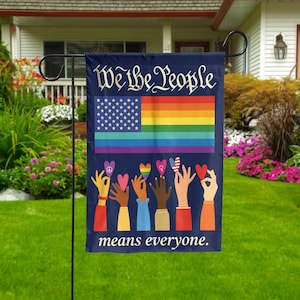 May include: A navy blue garden flag with the text "We the People" above a rainbow flag and American flag design. The phrase "means everyone" is printed below the flags, with hands holding hearts.