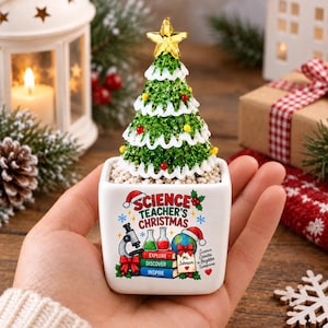 May include: A small white square ceramic planter containing a miniature green Christmas tree with white trim, colorful ornaments, and a gold star topper. The planter features the text "SCIENCE TEACHER'S CHRISTMAS" with graphics of a microscope, beakers, a globe, and books.