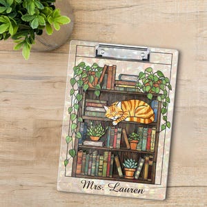 May include: A clipboard with a detailed illustration of a bookshelf with books, plants, and a sleeping orange tabby cat. The text "Mrs. Lauren" is printed at the bottom. The clipboard has a silver clip at the top.