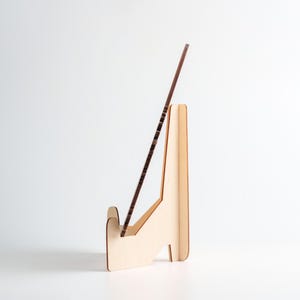 May include: A light-colored wooden display stand with a brown, rectangular object resting in its slot. The stand has a simple, angled design with a base for stability. The object appears to be a thin, framed piece.