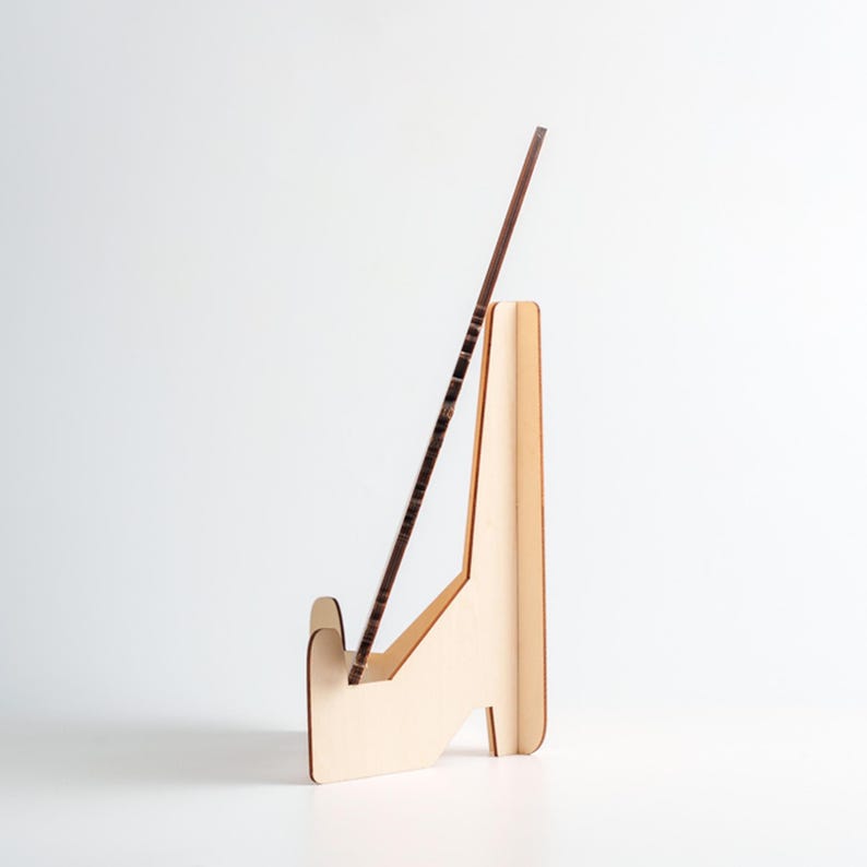 May include: A light-colored wooden display stand with a brown, rectangular object resting in its slot. The stand has a simple, angled design with a base for stability. The object appears to be a thin, framed piece.