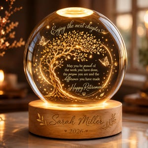 May include: A spherical glass ornament on a wooden base featuring an illuminated golden tree and butterfly design. The text reads: Enjoy the next chapter, May you be proud of the work you have done, the person you are and the difference you have made, Happy Retirement. The base is engraved with Sarah Miller and 2026.