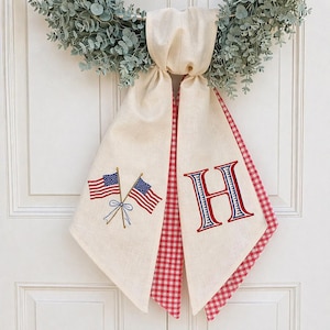 May include: Cream-colored fabric door hanger with embroidered American flags and the letter "H" in red and blue. The edges are trimmed with red and white gingham fabric. A decorative wreath hangs above.