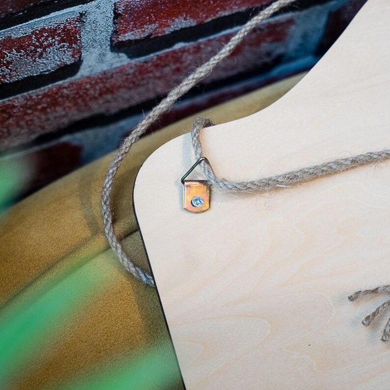 May include: Close-up of a wooden sign with a natural wood grain finish. A metal hanger is attached to the top corner with a gray rope for hanging. The background includes a brick wall and a glimpse of a painted surface.