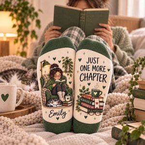 May include: A pair of cream-coloured socks with dark green cuffs and toes. The left sock features an illustration of a person reading in a green chair with a cat, labelled Emily. The right sock displays a stack of books, a mug, and the text Just One More Chapter.