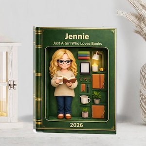 Op de afbeelding: Een groen, boekvormig decoratief item met de naam "Jennie" en de tekst "Just A Girl Who Loves Books". Binnenin is een 3D-figuur van een lezend meisje, met boeken, een bril en een koffiekopje. Het jaar "2026" staat onderaan.