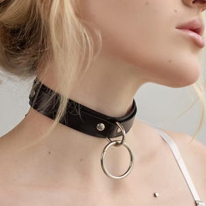 Pode incluir: Um colar gargantilha de couro preto com um pingente de anel de metal prateado. A gargantilha é presa com rebites prateados e um fecho de fivela. A modelo está usando um top de lingerie rosa e branco.
