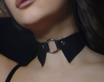 May include: Black choker necklace with a gold-colored ring and collar-style design. The choker has a sleek, minimalist aesthetic, with a focus on the circular metal accent. The collar is made of a matte black material.