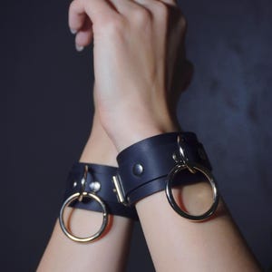 May include: Black leather wrist cuffs with large silver-colored metal rings. The cuffs are secured with metal snaps. The image shows two wrists with the cuffs, set against a dark background.