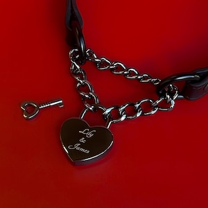 May include: A black leather collar with a silver chain and a heart-shaped padlock. The padlock is engraved with the names "Lily & James." A small key is next to the padlock. The background is a solid red.