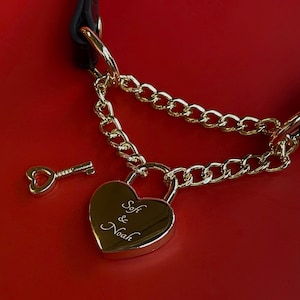 Może przedstawiać: Złote sercowate kłódka z wygrawerowanymi imionami "Sofi & Noah", połączona z łańcuchem i czarnym skórzanym paskiem. Obecny jest również klucz w kształcie serca. Tło jest jednolicie czerwone.