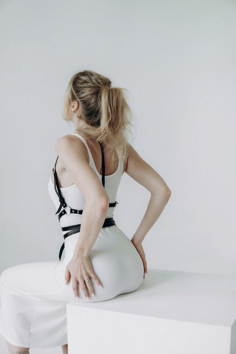 May include: A person wearing a white jumpsuit and a black leather harness. The person has blonde hair pulled back in a ponytail. The person is sitting on a white surface with their back to the camera.