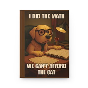 May include: A hardback journal featuring a cartoon dog wearing glasses, sitting at a desk. The cover text reads "I DID THE MATH, WE CAN'T AFFORD THE CAT."