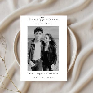 May include: A black and white Save the Date card featuring a couple walking on a beach. The card includes the names "Lola + Ben", the location "San Deigo, California", and the date "07.10.2025". The card is set on a white plate and a cream-colored fabric.