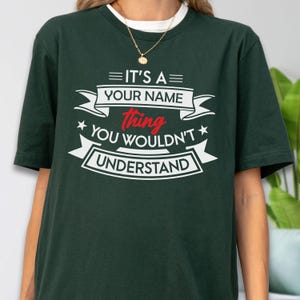 May include: A dark green short-sleeve t-shirt featuring white and red text that reads IT'S A YOUR NAME thing YOU WOULDN'T UNDERSTAND, arranged with decorative banners and stars on the chest. The shirt has a crew neckline and a relaxed fit.