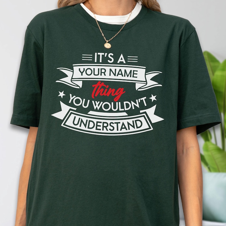 May include: A dark green short-sleeve t-shirt featuring white and red text that reads IT'S A YOUR NAME thing YOU WOULDN'T UNDERSTAND, arranged with decorative banners and stars on the chest. The shirt has a crew neckline and a relaxed fit.