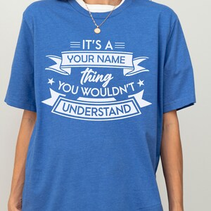 May include: A blue short-sleeved t-shirt featuring white screen-printed text that reads IT'S A YOUR NAME thing YOU WOULDN'T UNDERSTAND, arranged within decorative banners and star accents. The shirt has a crew neckline and a heathered fabric texture.