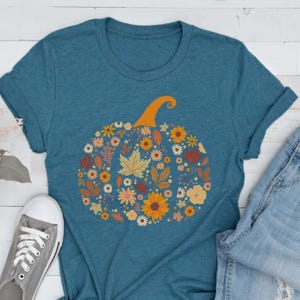 May include: Teal-colored t-shirt featuring a floral pumpkin design. The pumpkin is composed of various flowers and autumn leaves in shades of orange, yellow, and brown. The shirt has short sleeves and a crew neck, with a pair of jeans and a gray sneaker visible.