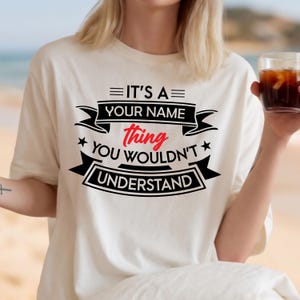May include: A cream-colored short-sleeve t-shirt featuring black and red text that reads IT'S A YOUR NAME thing YOU WOULDN'T UNDERSTAND. The design includes black banners, stars, and horizontal lines surrounding the bold, centered lettering.