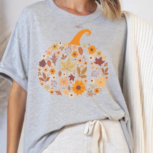 May include: A light gray t-shirt featuring a floral pumpkin design. The pumpkin is composed of various autumn-colored flowers and leaves, with an orange stem. The shirt is a short-sleeved crew neck.