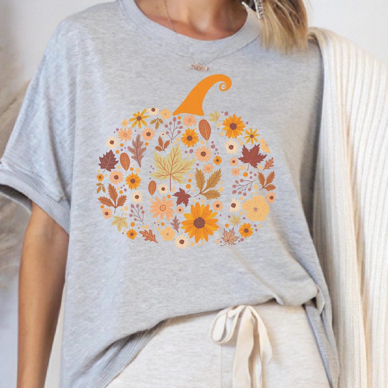 May include: A light gray t-shirt featuring a floral pumpkin design. The pumpkin is composed of various autumn-colored flowers and leaves, with an orange stem. The shirt is a short-sleeved crew neck.