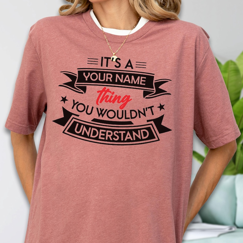 May include: A mauve short-sleeved t-shirt featuring black and red text that reads: IT'S A YOUR NAME thing YOU WOULDN'T UNDERSTAND. The design includes decorative black banners and stars surrounding the red word thing on the front of the shirt.