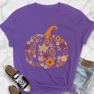 May include: A purple t-shirt featuring a floral pumpkin design. The pumpkin is composed of various flowers and leaves in shades of orange, yellow, and brown, with an orange stem. The shirt is paired with black sneakers and blue jeans.