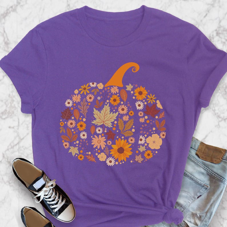 May include: A purple t-shirt featuring a floral pumpkin design. The pumpkin is composed of various flowers and leaves in shades of orange, yellow, and brown, with an orange stem. The shirt is paired with black sneakers and blue jeans.