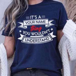 May include: A navy blue short-sleeved t-shirt featuring white and red graphic text that reads IT'S A YOUR NAME thing YOU WOULDN'T UNDERSTAND, arranged with decorative banners and stars across the chest area of the garment.