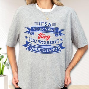 May include: A heather gray short-sleeve t-shirt featuring blue and red text that reads IT'S A YOUR NAME thing YOU WOULDN'T UNDERSTAND, arranged with blue banners and stars. The shirt has a crew neckline and a simple, casual cut for everyday wear.
