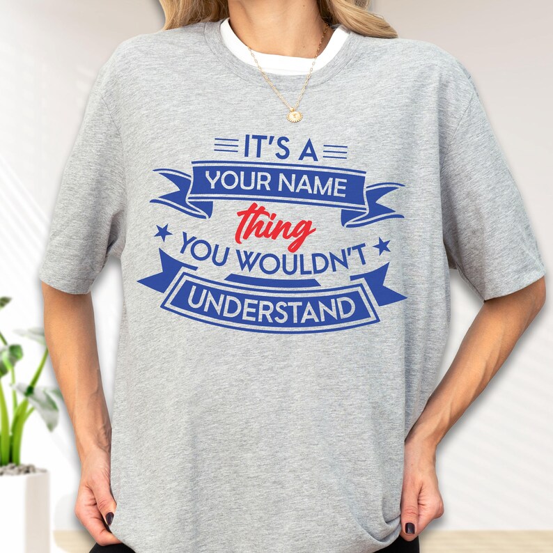 May include: A heather gray short-sleeve t-shirt featuring blue and red text that reads IT'S A YOUR NAME thing YOU WOULDN'T UNDERSTAND, arranged with blue banners and stars. The shirt has a crew neckline and a simple, casual cut for everyday wear.